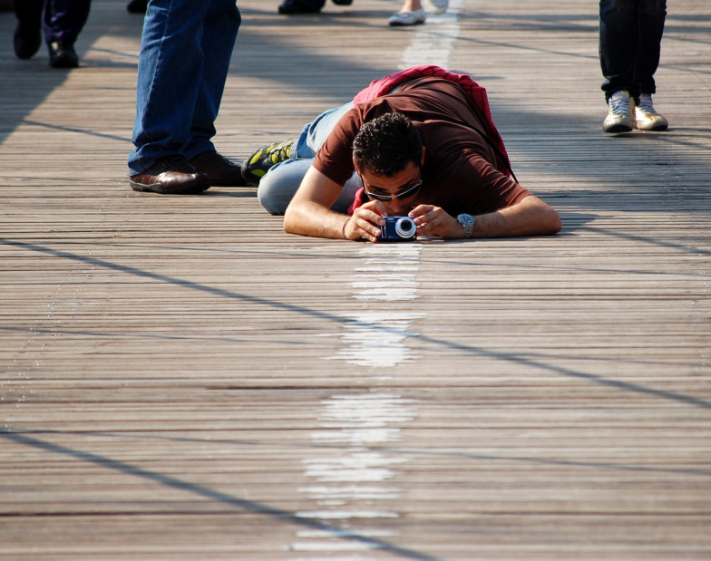 Foto: ein Mann mit Fotoapparat liegt zum Fotografieren auf den Holzplanken einer Brücke