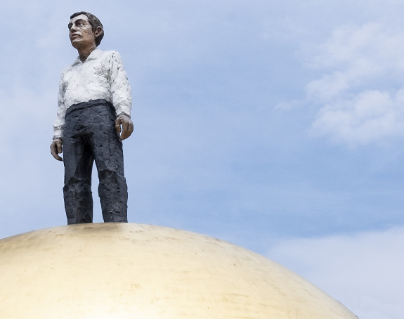 Photo of a sculpture: A man in a white shirt and black trousers stands on a golden ball