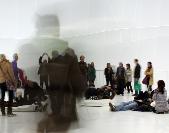Foto: auf einer Ausstellungsfläche halten sich Personen auf - stehend und liegend. Das Bild wurde mit einer weiteren stehenden Person überblendet.