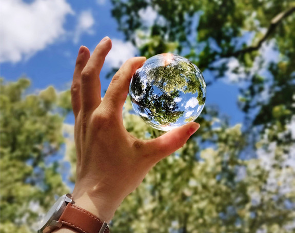 Das Foto zeigt eine Hand, die zwischen Daumen und Zeigefinger einer Glaskugel in Richtung Himmel und Baumkronen hält.