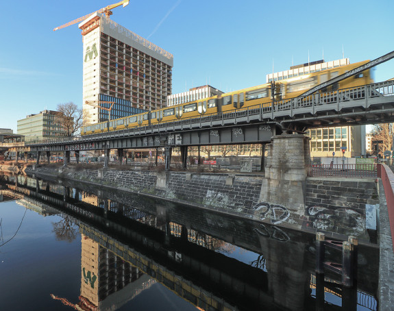 U-Bahn fährt über Brücke mit Kränen im Hintergrund.